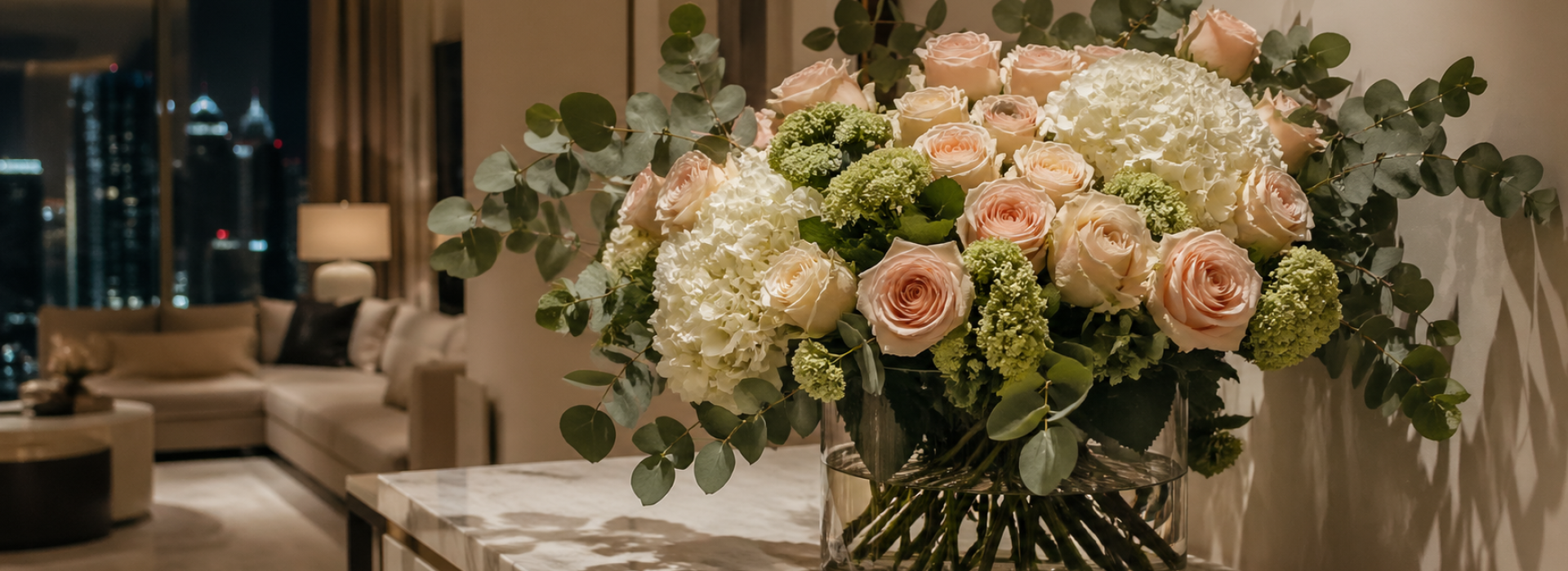 Flower styling Dubai home — blush roses and white hydrangeas in a glass vase on a marble console with Dubai city lights at night